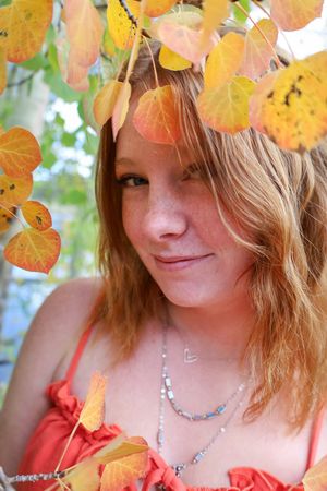 Young redhead woman with freckles smiling among vibrant orange autumn leaves wearing a coral dress and silver necklace.