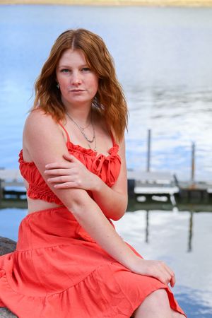 Young redhead woman in coral dress sitting on rocks by a calm lake with a dock visible in background.