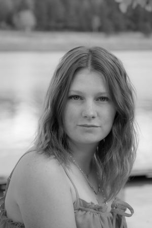 Black and white portrait of a young woman with long hair wearing a sundress near a reflective lake.