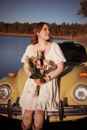 Young woman in white dress holding flowers leaning against a vintage yellow car by a lake.