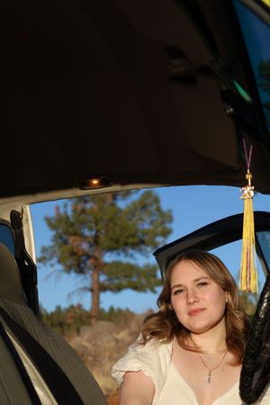 Young woman in a white dress sitting inside a car with a graduation tassel hanging visible.
