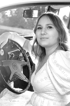 Young woman in white dress posing in vintage car, black and white portrait photo.