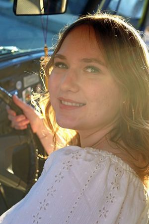 Young woman smiling in car with golden sunlight streaming through window, wearing white dress.