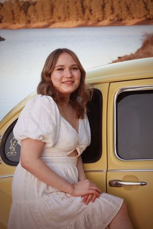 Smiling young woman in white eyelet dress leaning against vintage yellow VW Beetle by lake.