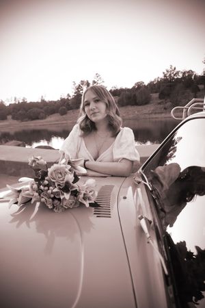 Young woman in white dress holding bouquet leaning on vintage car hood, sepia tone photo.