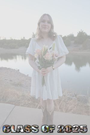 Class of 2025 senior girl in white dress holding pink tulips standing by peaceful lake.