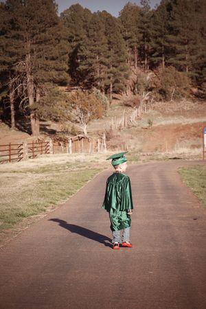 Young boy in green graduation cap and gown standing alone on rural driveway among pine trees.