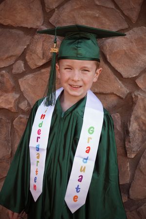 Happy kindergarten graduate boy smiling in green cap and gown with colorful graduation stole.