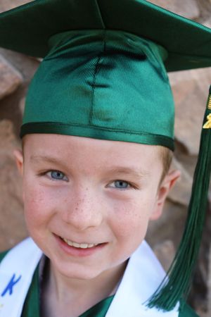 Smiling freckled boy with blue eyes wearing green kindergarten graduation cap and gown.