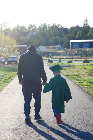 Adult and young child in green graduation cap and gown walking hand in hand down a sunny path.