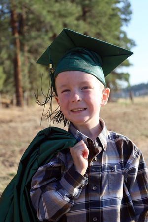 Smiling boy in green graduation cap and gown holding robe over shoulder outdoors among pine trees.