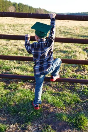 Young boy in green graduation cap climbing a rustic metal fence in a rural field setting.