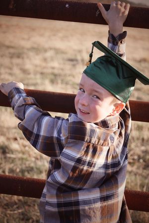 Smiling boy in green graduation cap climbing a rustic metal fence with a sunny field background.