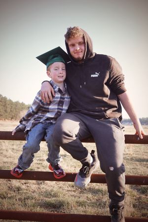 Young boy in green graduation cap sitting on a fence with an older teen in a Puma hoodie.