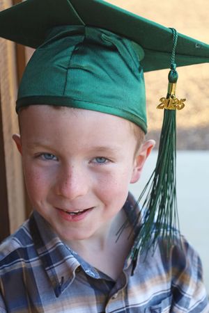 Close-up of a freckled boy smiling in a green 2025 graduation cap with gold tassel outdoors.