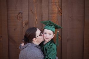 Mother kissing her young son wearing a green graduation cap and gown against a wooden fence.
