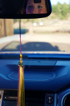 Gold and purple graduation tassel hanging from a car rearview mirror with a child reflected above.