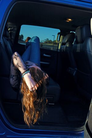 Young woman lying upside down in back seat of blue truck, wearing jeans and black lace top at sunset.