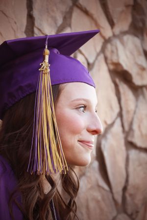 Young woman in purple graduation cap and gown smiling in profile against stone wall background.
