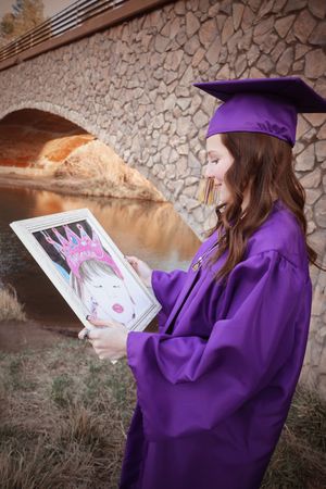 Graduate in purple cap and gown holding framed childhood photo near stone bridge at sunset.