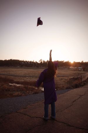 Graduate in purple gown tossing cap into air at sunset on rural road, silhouetted.