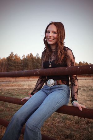 Smiling young woman in black lace top and jeans sitting on rustic fence rail at dusk.