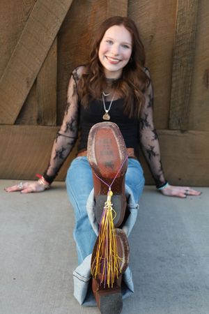 Smiling young woman in black lace top and jeans sitting with cowboy boot and graduation tassel forward.