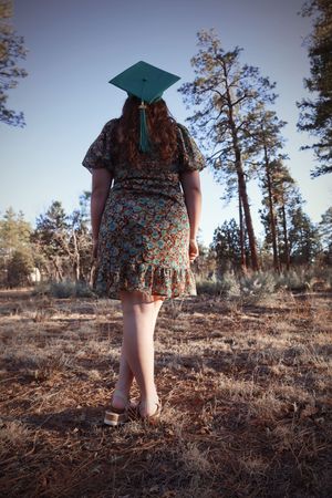 Young woman in floral dress and teal graduation cap walking through a pine forest.