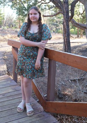 Smiling young girl in floral dress posing on a wooden bridge in a natural outdoor setting.