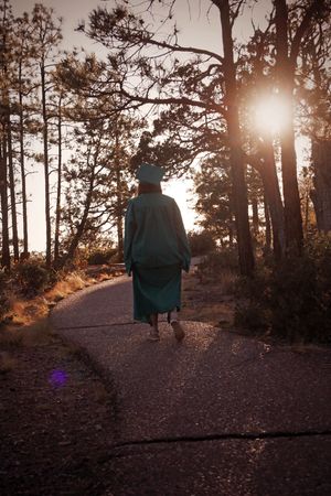 Graduate in teal cap and gown walking along a forest path toward a glowing sunset.