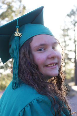 Smiling young graduate in teal cap and gown with 2025 tassel charm outdoors in sunlight.
