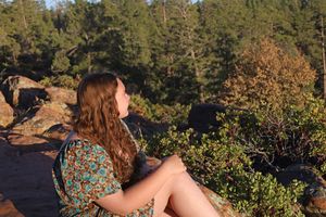 Young woman in floral dress sitting on rocks gazing at a lush green forest landscape.