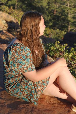 Young woman with long curly hair in floral dress sitting on a rock in golden sunlight.