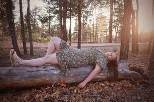 Young woman in floral dress lying on a fallen log in a sunlit pine forest during golden hour.