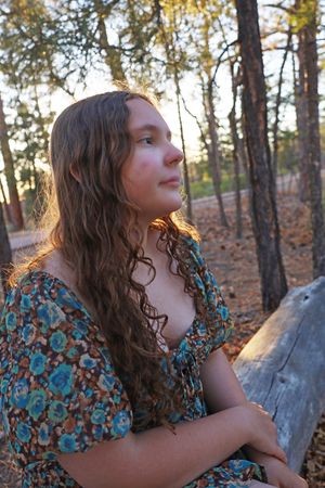 Young woman with long curly hair in floral dress sitting outdoors in warm golden forest light.