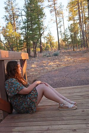 Young woman in floral dress sitting on wooden deck leaning against bench surrounded by pine forest at sunset.