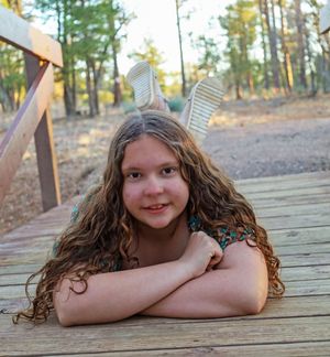 Smiling girl with long curly hair lying on wooden deck in a pine forest setting.