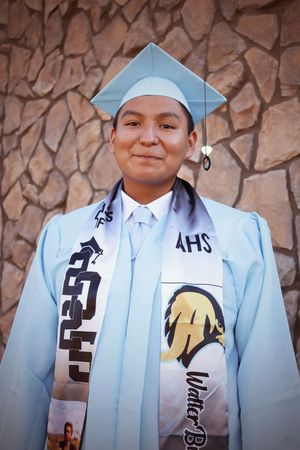 Young male graduate smiling in light blue cap and gown with AHS stole against stone wall.