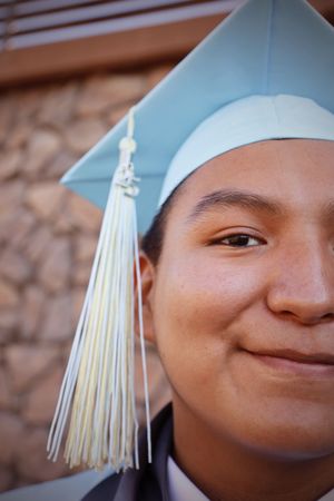 Half-face close-up of smiling graduate in light blue cap with white tassel against stone wall.