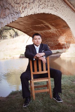 Young man in black suit sitting on wooden chair under stone arch bridge near river.