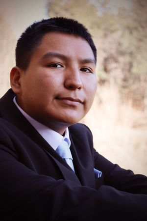 Young man in black suit and light blue tie posing with arms crossed in outdoor setting.