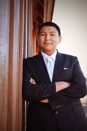Young man in black suit and blue tie standing with arms crossed against wooden column.