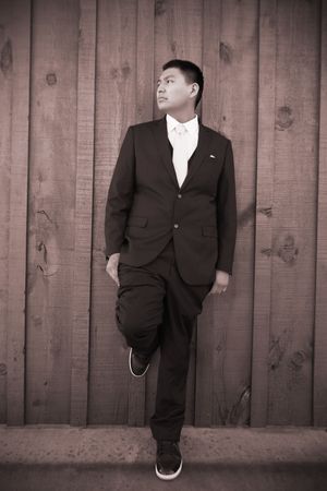 Young Asian man in black suit leaning against wooden wall with one foot raised, sepia tone portrait.