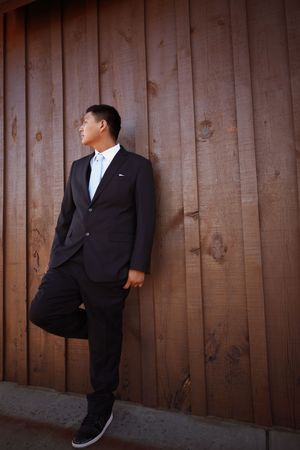 Stylish young Asian man in dark suit leaning against rustic wooden wall with leg raised outdoors.