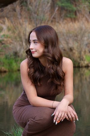 Beautiful young woman with long wavy brown hair in brown outfit sitting near a reflective pond.