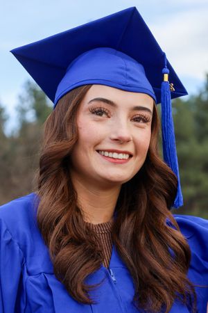 Smiling young woman in blue graduation cap and gown with long brown hair against sky background.