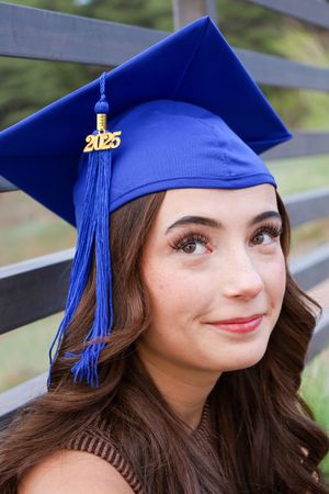 Close-up portrait of young woman in blue 2025 graduation cap with long brown wavy hair smiling.