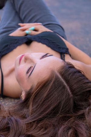 Young woman with long brown hair lying on ground with eyes closed wearing gray crop top outdoors.