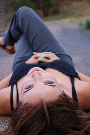 Young woman with brown hair lying on pavement looking up at camera wearing gray outfit outdoors.