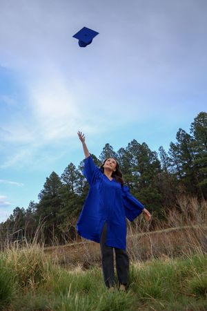 Graduate in blue cap and gown tossing mortarboard into sky in grassy field with pine trees behind.